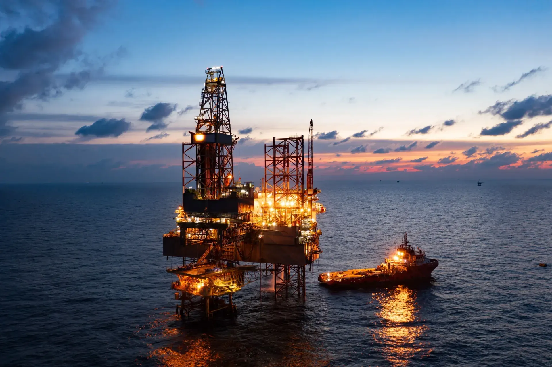 Offshore jack-up drilling rig and supply vessel at sea under a dusk sky