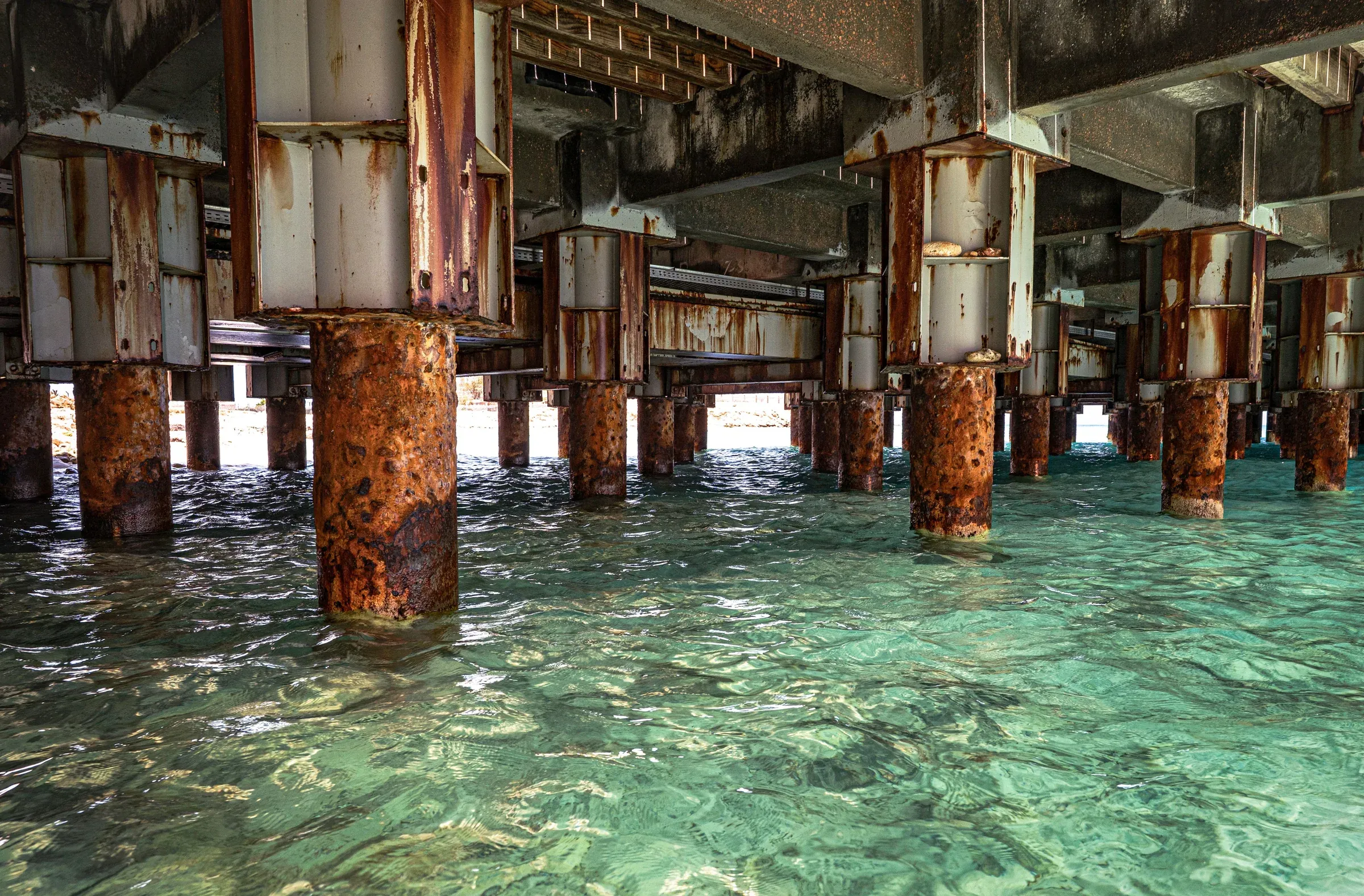Corroded steel pilings supporting a pier deck above clear shallow seawater